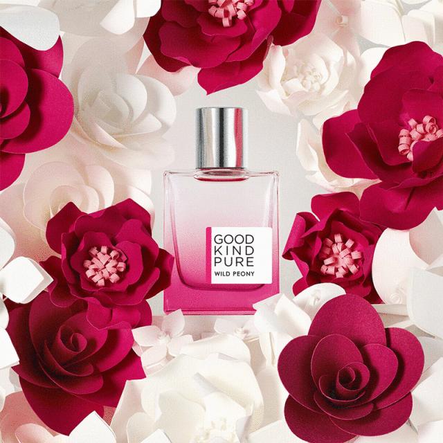 Close-up of a "Good Kind Pure Wild Peony" perfume bottle with a silver cap, surrounded by red and white paper flowers.