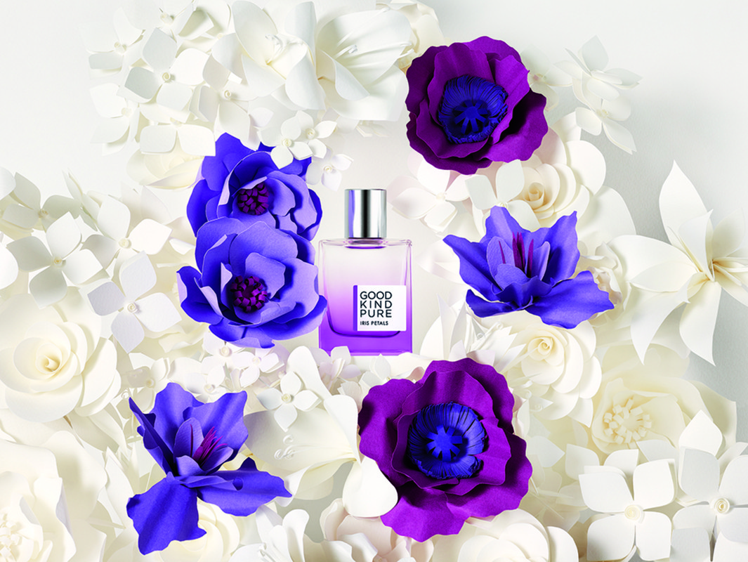 Perfume bottle centered, surrounded by white and purple paper flowers on a white background, showcasing a beauty and fragrance advertisement.