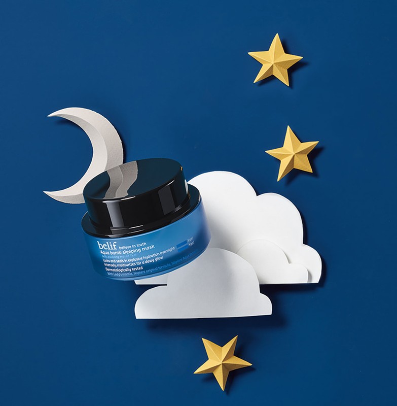 A blue jar of Belif Aqua Bomb sleeping mask is displayed with paper stars, clouds, and a crescent moon against a blue background.