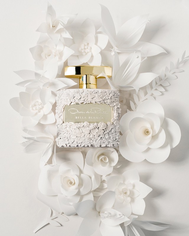A perfume bottle with a gold cap sits centered amongst white paper flowers in a fragrance ad, creating an elegant and floral composition.