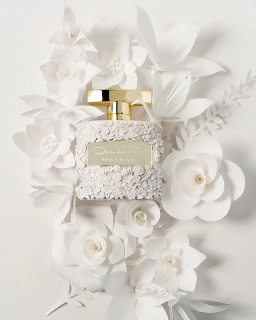 A perfume bottle with a gold cap sits centered amongst white paper flowers in a fragrance ad, creating an elegant and floral composition.