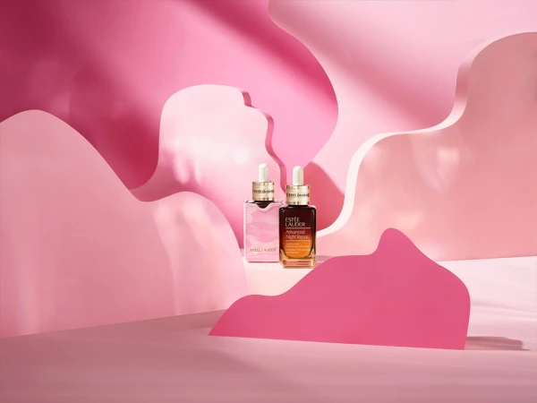 Two Estee Lauder Advanced Night Repair serum bottles displayed in front of pink paper cutouts for a beauty product advertisement.