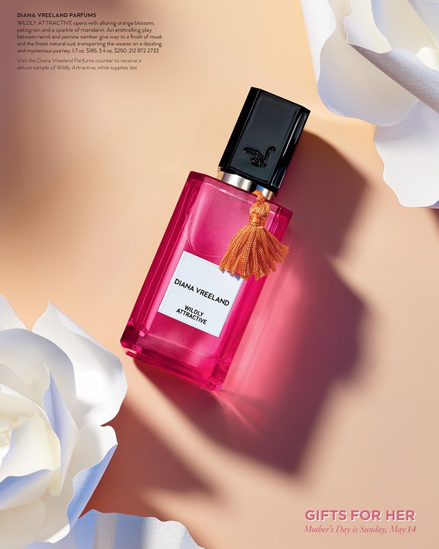 Rectangular pink perfume bottle with gold tassel and white flowers against a peach background in a beauty product photography style.