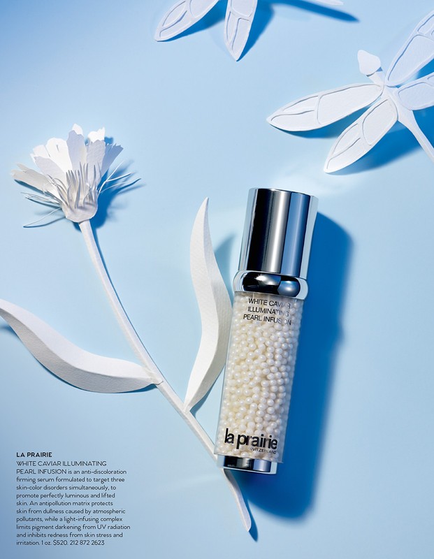 La Prairie White Caviar Illuminating Pearl Infusion serum in a clear and silver bottle, displayed with white paper flowers and dragonflies.