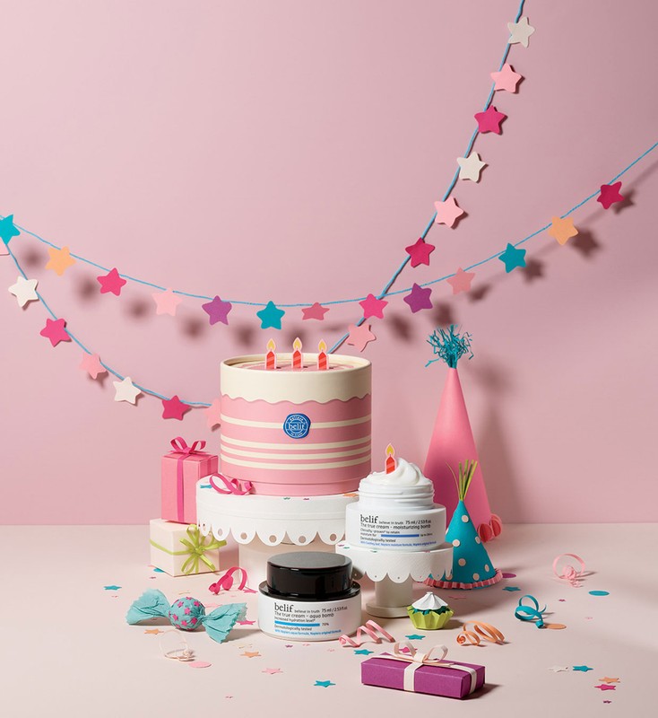 A whimsically styled beauty product shoot features skincare items arranged with a cake, gifts, and party decorations on a pink backdrop.