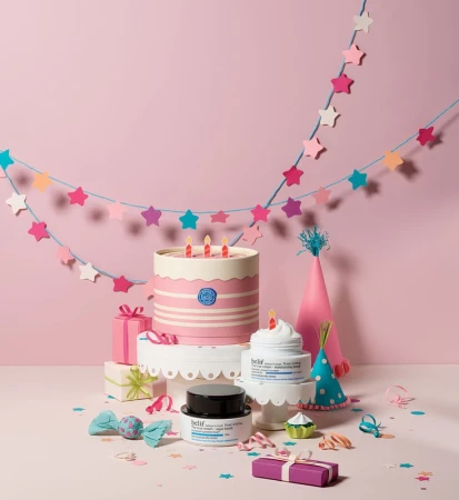 A whimsically styled beauty product shoot features skincare items arranged with a cake, gifts, and party decorations on a pink backdrop.
