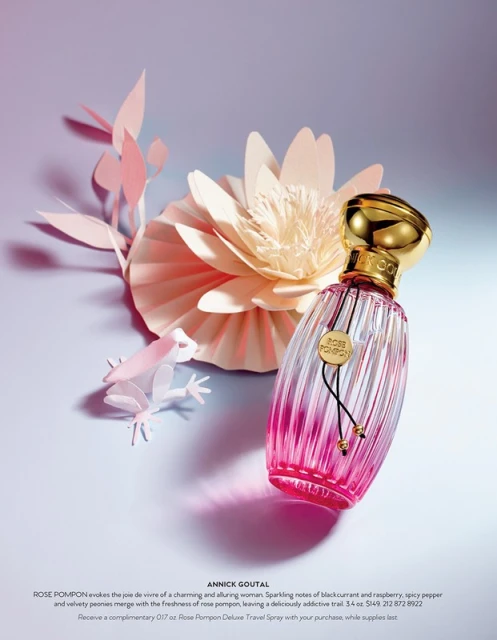 Perfume bottle with pink ombre effect and gold cap sits near paper flower art, creating an elegant beauty product still life.