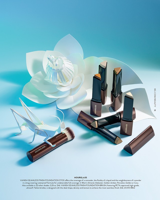 Hourglass makeup products, including foundation sticks and a brush, are arranged against a light blue backdrop with a paper flower.