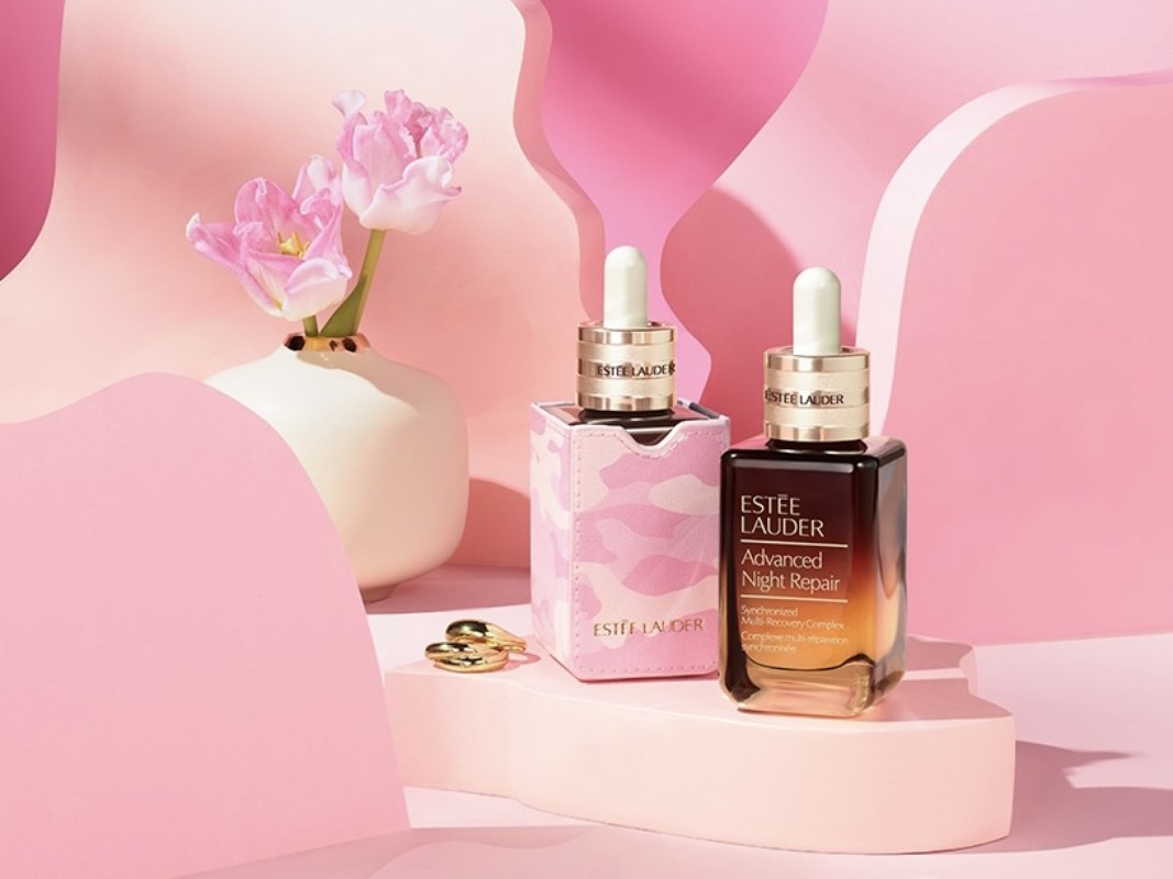 Estée Lauder Advanced Night Repair serum bottles on a pink platform with tulips in a white vase against a pink backdrop.