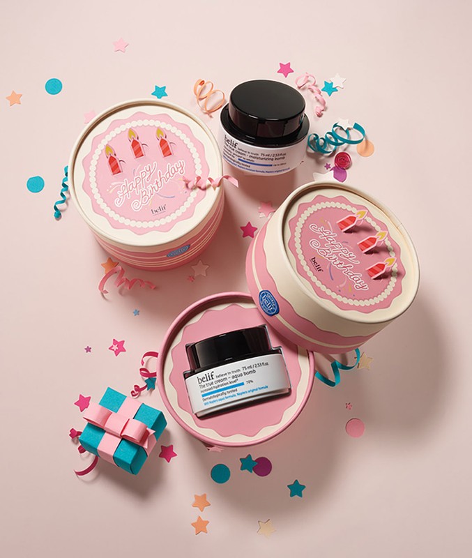 Belif skincare product assortment on a pink background with confetti and a wrapped gift, in cake-themed packaging for a birthday promotion.