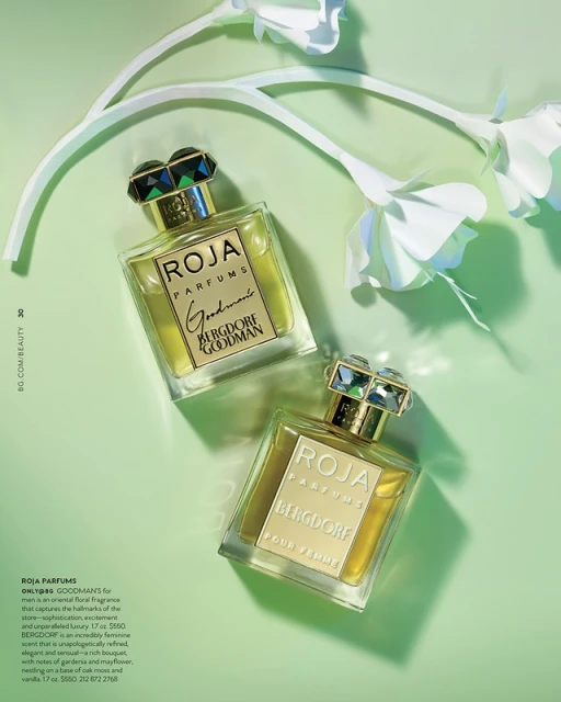 Two Roja Parfums fragrance bottles with faceted caps on a soft green background, accented by white paper flower decorations.