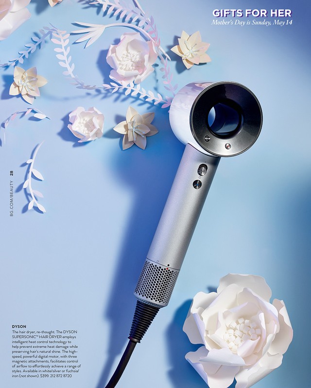 Studio shot of a Dyson Supersonic hair dryer against a light blue backdrop, accented with white paper flowers and leafy paper cutouts.