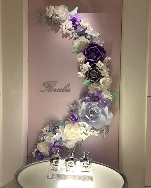 Floral display with paper flowers in white, purple, and grey, accompanied by three perfume bottles on a clear pedestal, artful composition.