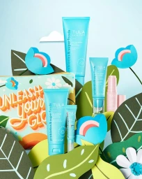 Tula Skincare tubes and cosmetic pouch arranged among paper art tropical leaves and blue flowers on a light blue background.