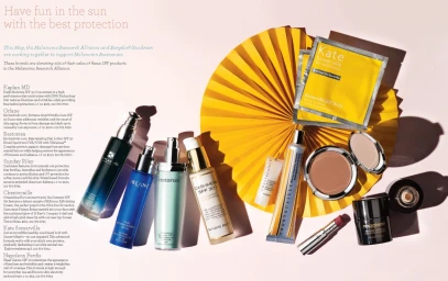 Flat lay of SPF skincare products and makeup arranged on a pale surface next to a yellow folded paper fan.