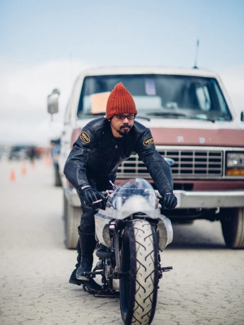 Man wearing a beanie and leather jacket is riding a motorcycle on a dry lake bed. Vintage van and other people are in the background.