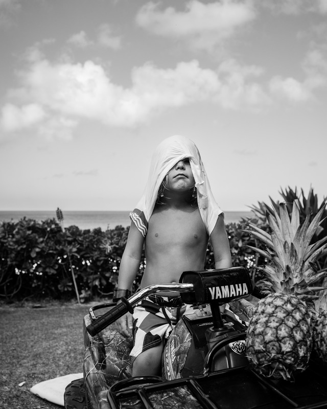 A young child sits on a Yamaha ATV next to pineapples with a white cloth draped over their head, ocean and cloudy sky behind.