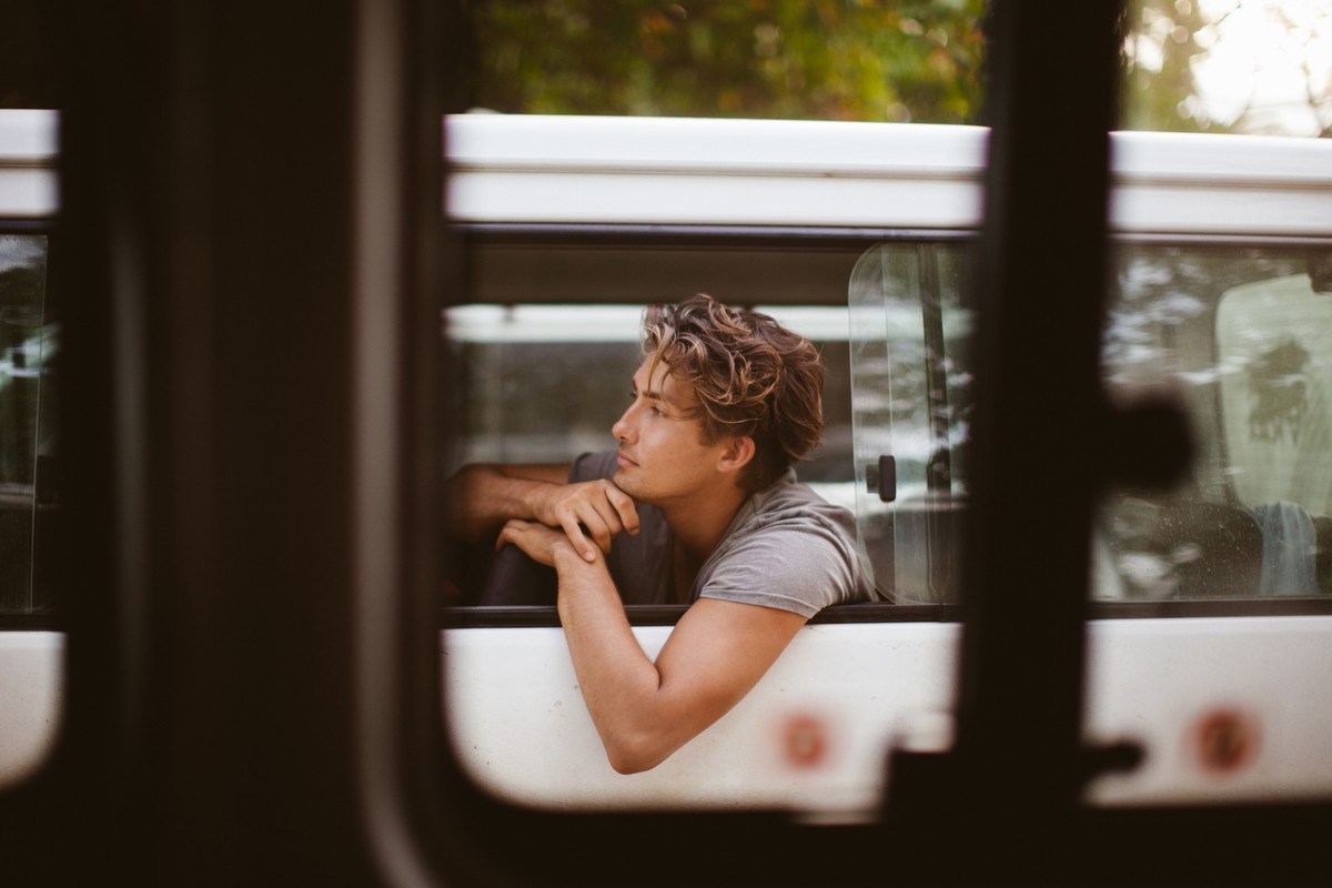 A young man with curly brown hair sits inside a white bus, his chin resting on his hands as he gazes out the window.