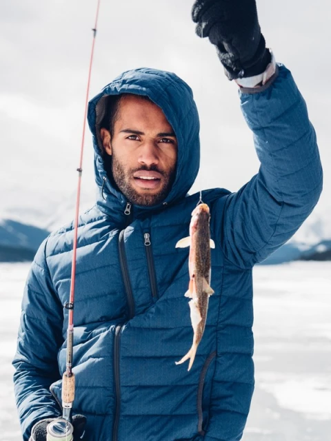Man ice fishing in a blue hooded jacket, holding a caught fish up on the fishing line with snowy mountains in the background.