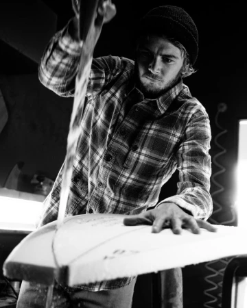 Monochrome portrait of a man wearing a beanie and flannel shirt, shaping a surfboard with a tool, in a workshop setting with dark background.