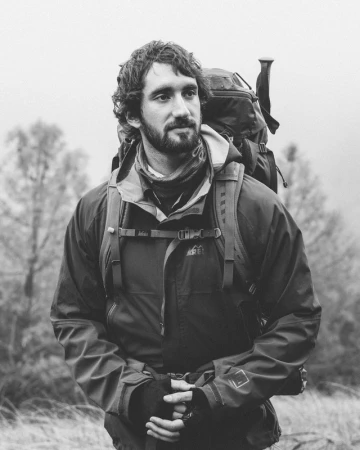 Monochrome portrait of a bearded man wearing a hiking jacket and carrying a heavy backpack outdoors
