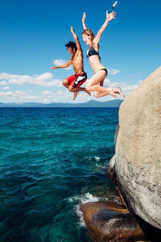 A man and a woman jump off a rock into the ocean against a blue sky with a sunny and bright aesthetic, conveying travel and fitness.