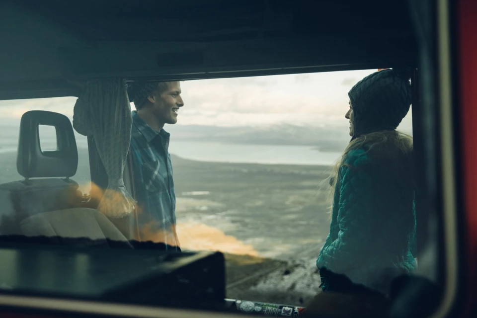 A young couple in a camper van enjoys a scenic view. One man, one woman, smiles in a bright outdoor landscape with diffused lighting.