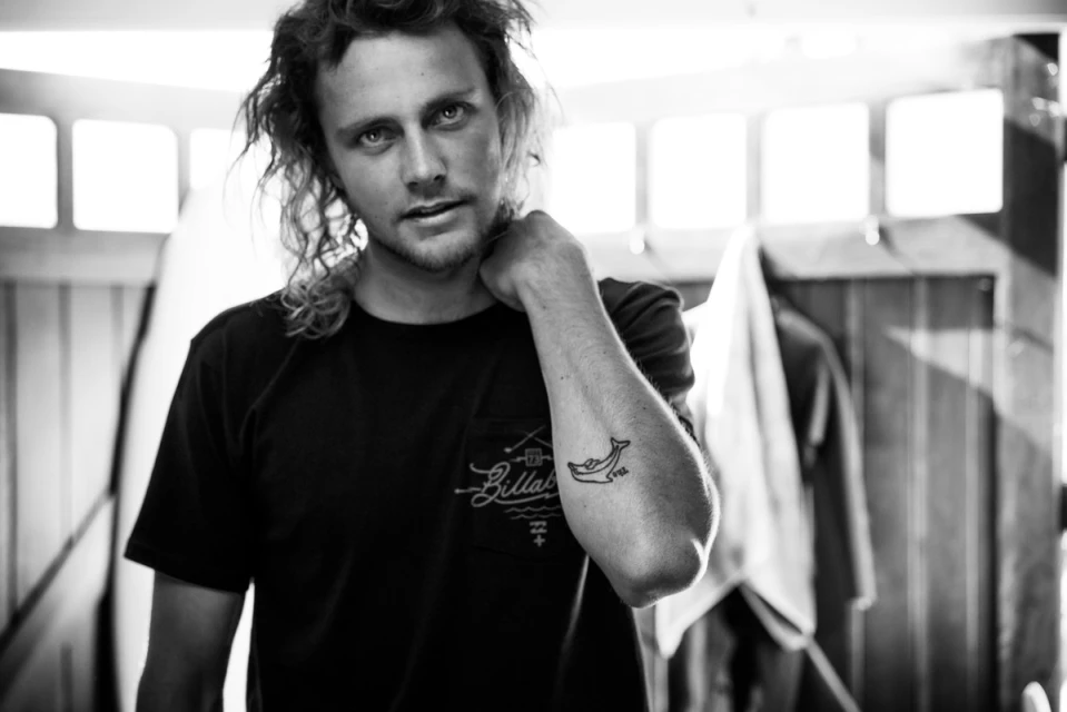 Black and white portrait of a young man with curly hair, wearing a t-shirt with a logo and a dolphin tattoo on his arm.