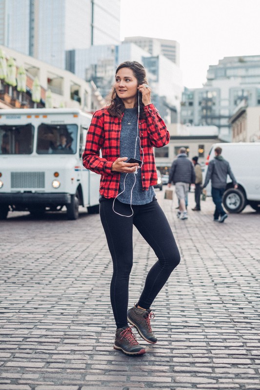 Woman in red plaid listens to headphones on city street with a food truck and buildings in the background, holding a smartphone.