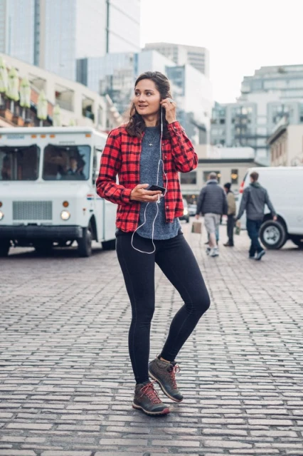 Woman in red plaid listens to headphones on city street with a food truck and buildings in the background, holding a smartphone.