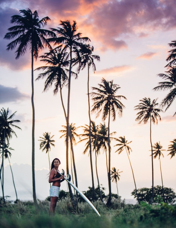Woman standing on a grassy field holding a surfboard against a colorful sunset sky with palm trees in the background on a tropical location.