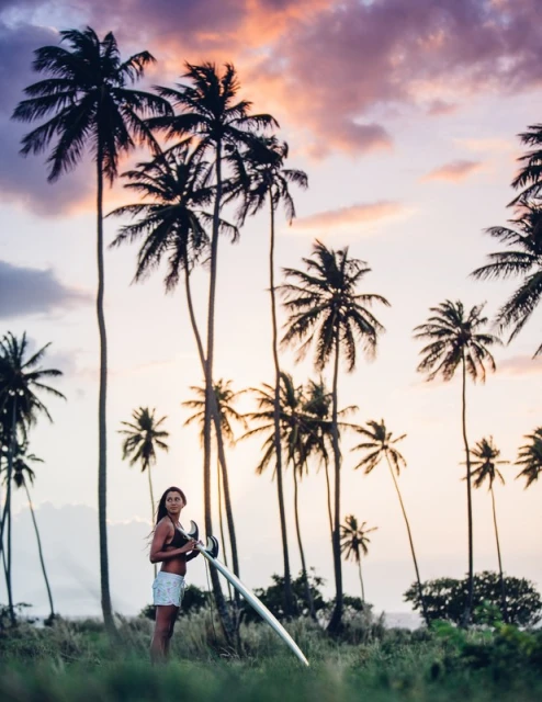 Woman standing on a grassy field holding a surfboard against a colorful sunset sky with palm trees in the background on a tropical location.
