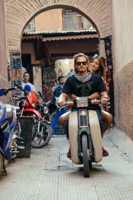 Couple riding a vintage moped through a narrow, sunlit street framed by an archway, capturing urban travel and retro style.
