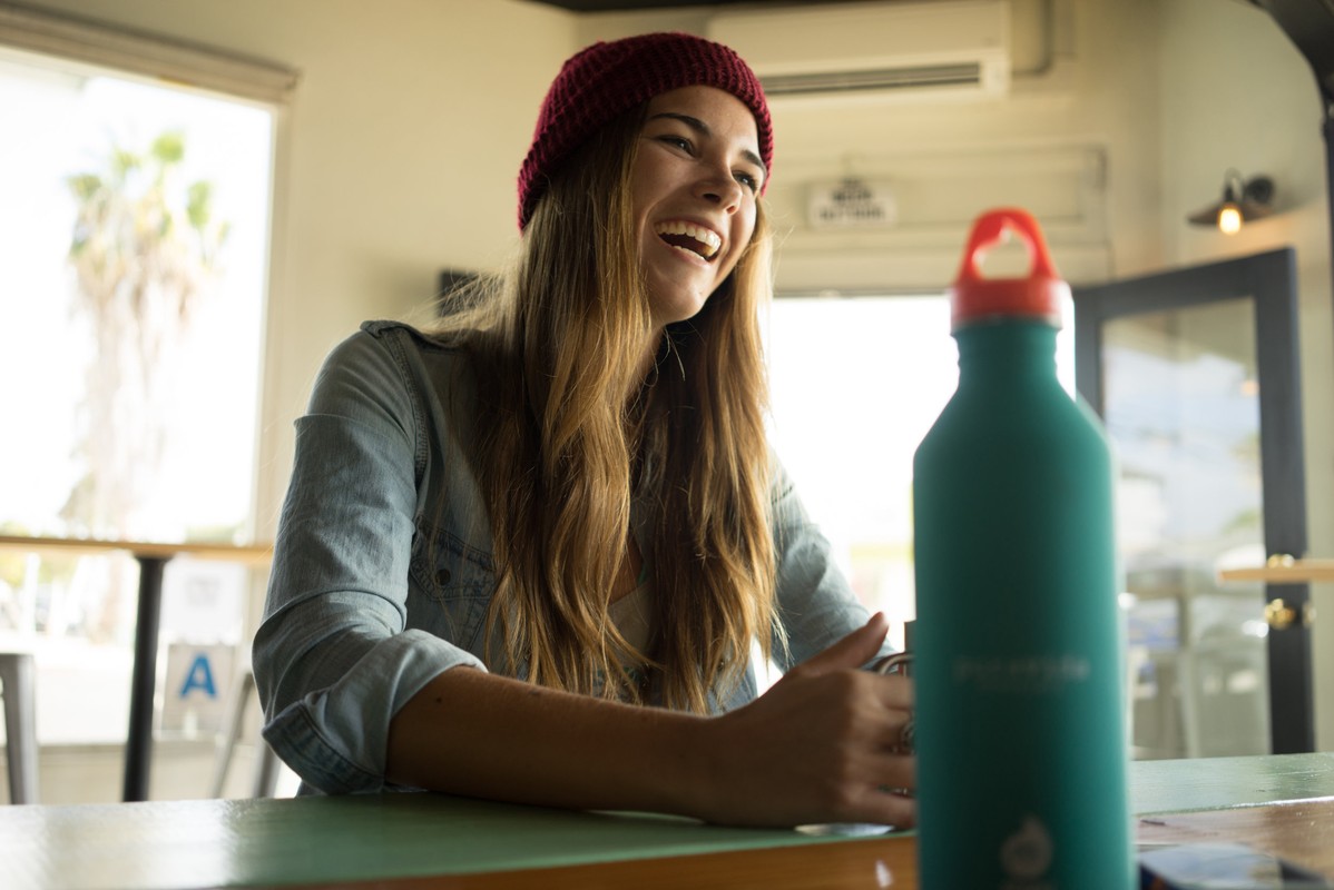 Young woman with red beanie laughs at a table with a teal water bottle in an indoor setting with a window and natural light.