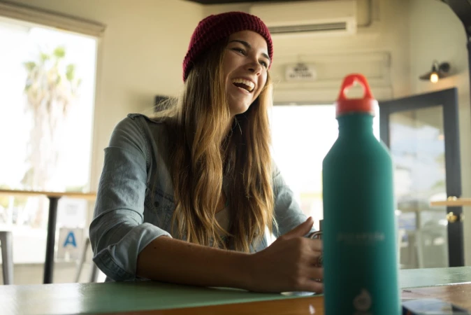 Young woman with red beanie laughs at a table with a teal water bottle in an indoor setting with a window and natural light.