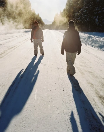 Two people walk on a snow-covered road lined with trees under bright sunlight, casting long shadows, with misty air in the background.