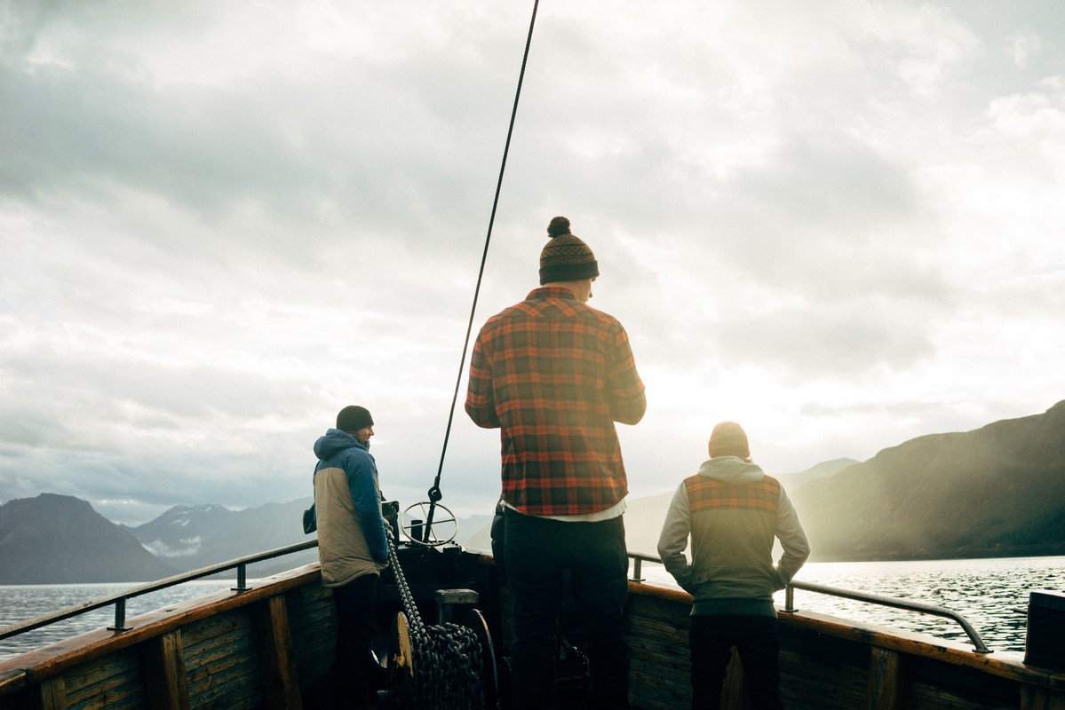 Three people aboard a wooden boat on a lake against a cloudy sky and distant mountains, creating a serene outdoor adventure scene.