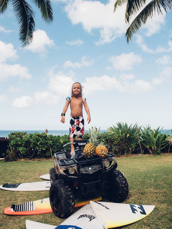 A shirtless young boy stands on a four-wheeler with pineapples on a lawn with surfboards and the ocean visible in the background.
