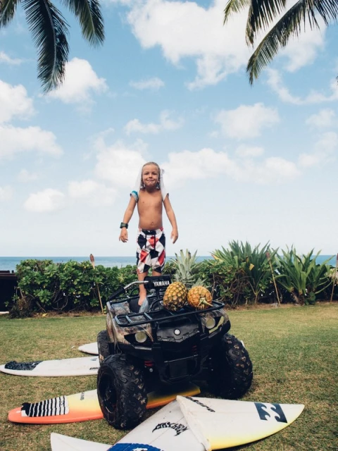 A shirtless young boy stands on a four-wheeler with pineapples on a lawn with surfboards and the ocean visible in the background.