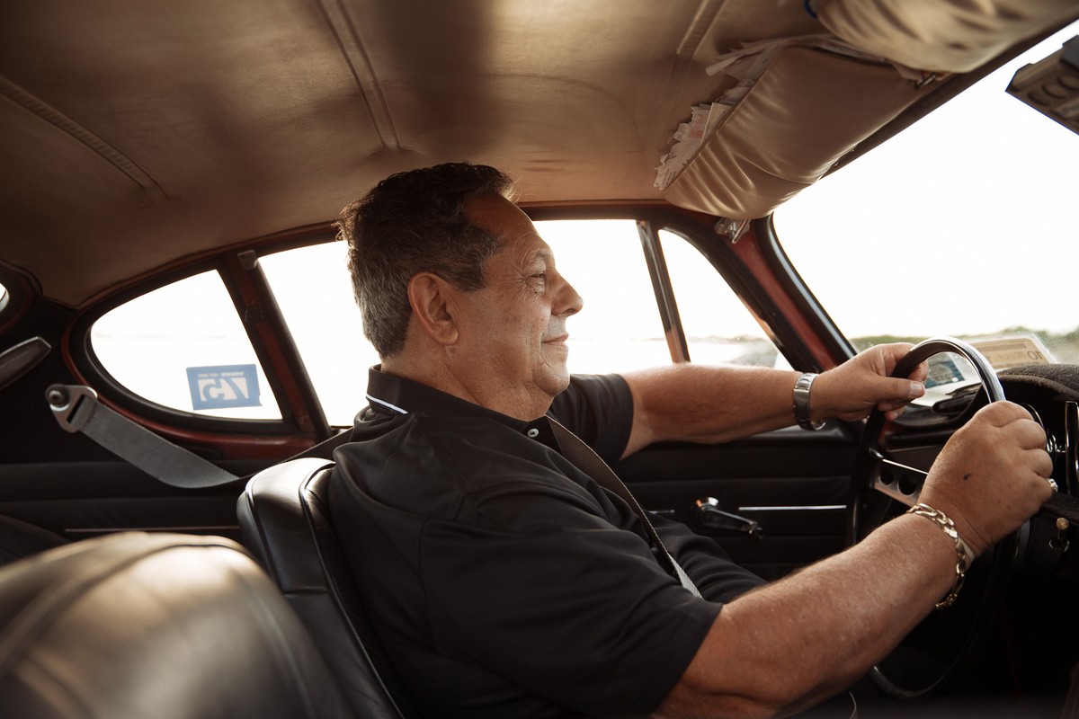 Man driving classic car with black interior. Wearing a black collared shirt, he grips the steering wheel, looking out the window.