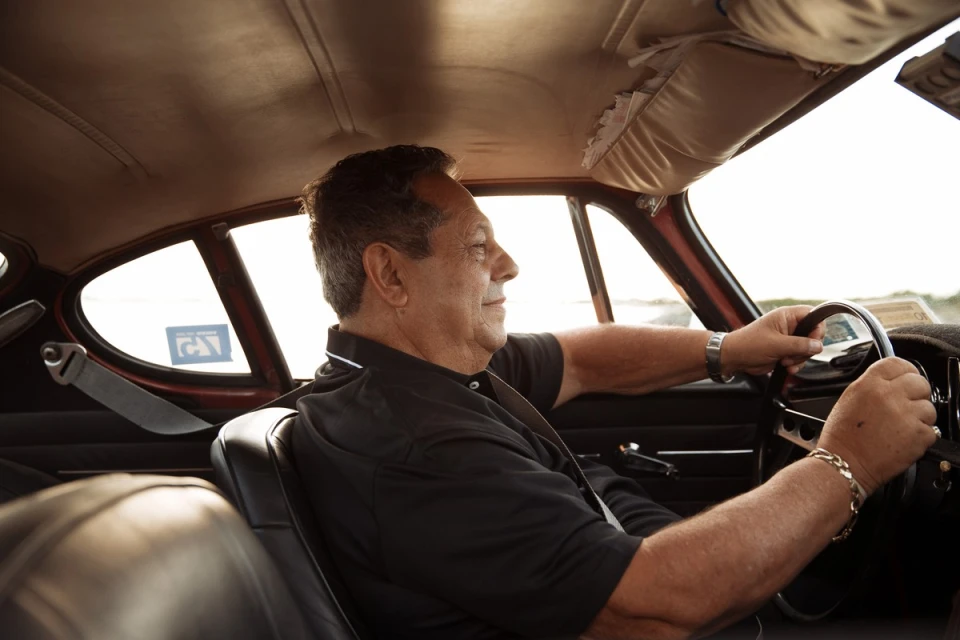 Man driving classic car with black interior. Wearing a black collared shirt, he grips the steering wheel, looking out the window.