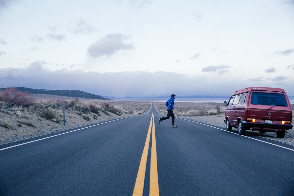 Adult male crossing a long asphalt road on foot, rural landscape with a parked van, cloudy sky and distant hills.