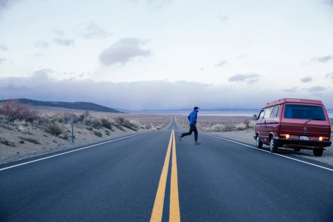Adult male crossing a long asphalt road on foot, rural landscape with a parked van, cloudy sky and distant hills.