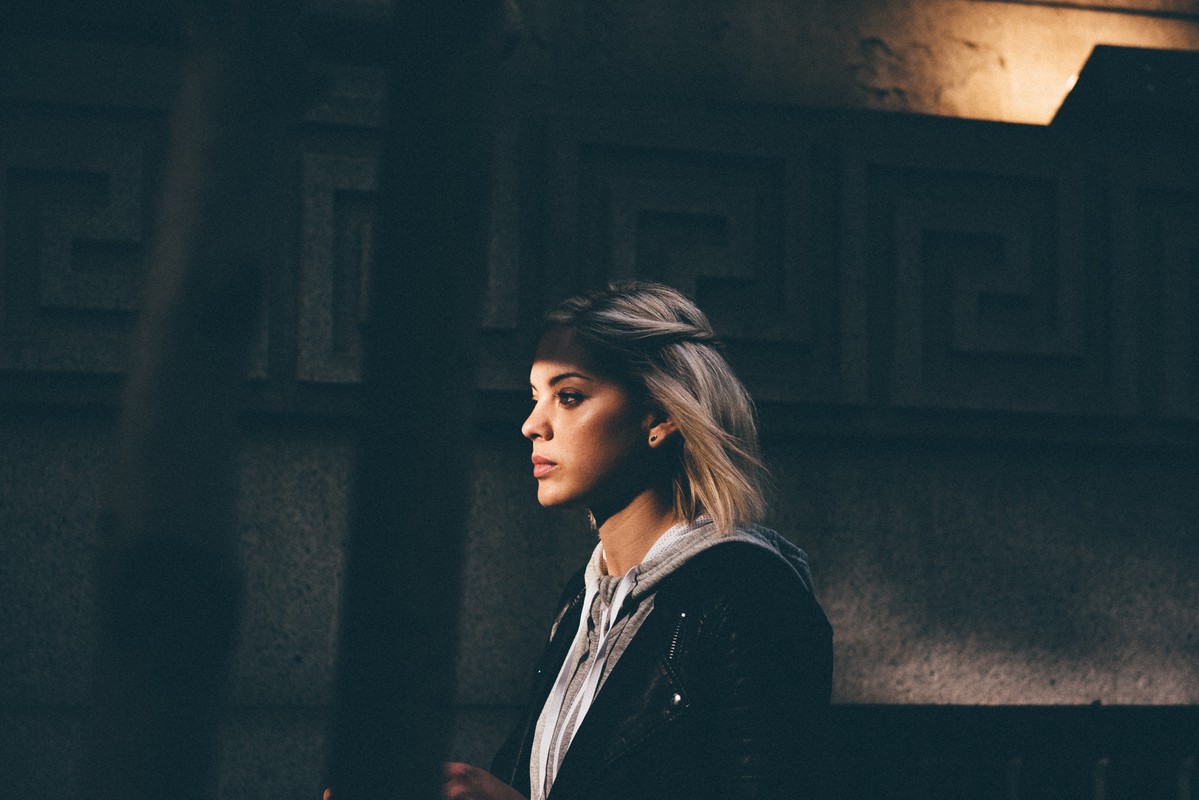 Portrait of a young woman with blonde hair in an urban setting, wearing a hooded sweatshirt and leather jacket in a dimly lit outdoor environment.