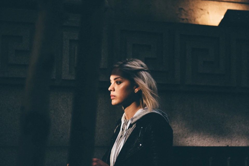 Portrait of a young woman with blonde hair in an urban setting, wearing a hooded sweatshirt and leather jacket in a dimly lit outdoor environment.