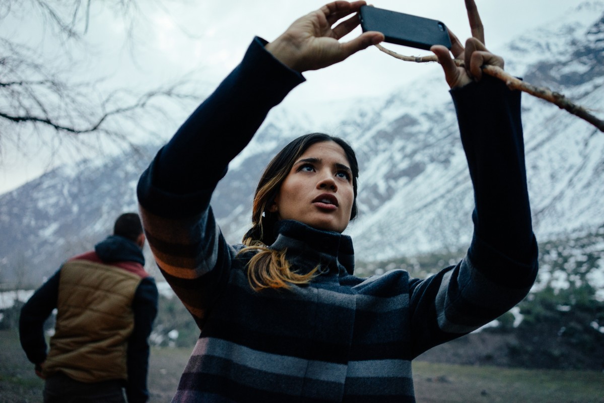 Woman outdoors taking photo with phone, another person in background, snow-capped mountains, neutral lighting, medium shot.