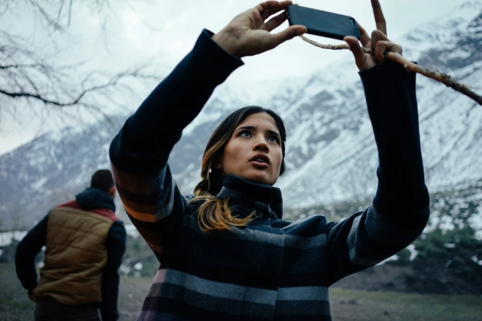 Woman outdoors taking photo with phone, another person in background, snow-capped mountains, neutral lighting, medium shot.