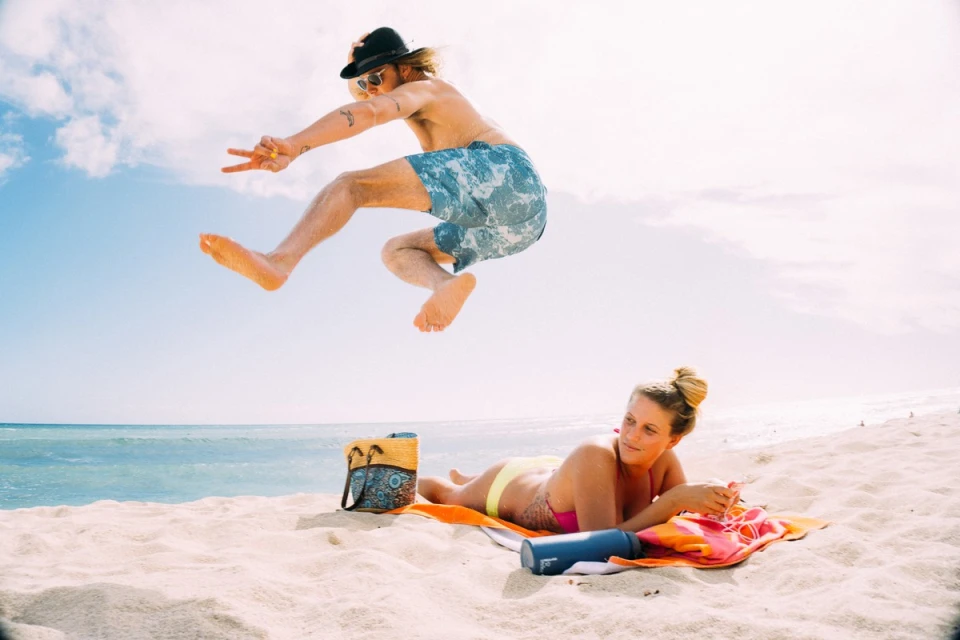 Man jumping on a beach near woman sunbathing, clear blue sky and ocean background, a sunny day with bright, natural lighting.