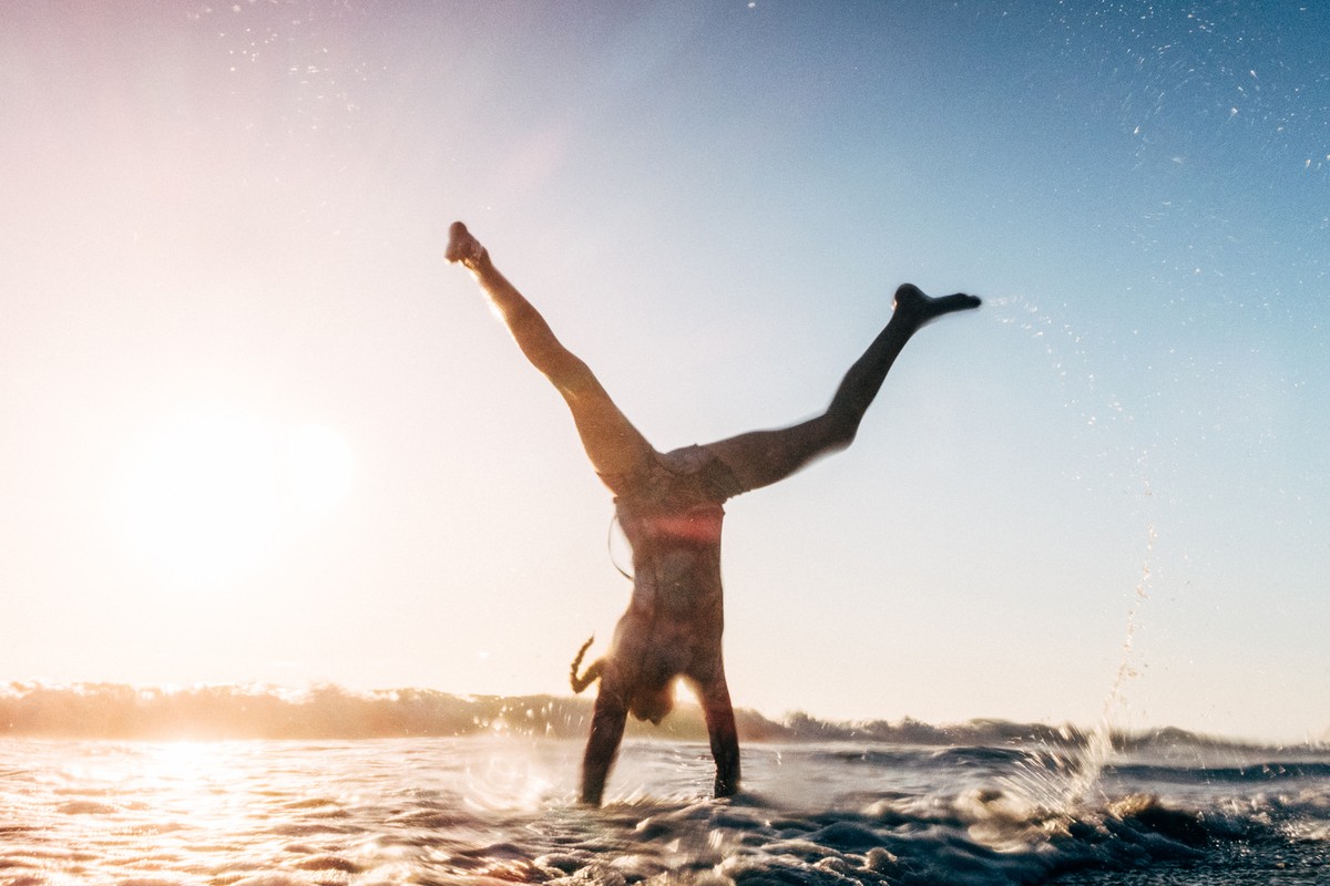 A person performs a handstand on a beach as waves roll in, illuminated by the sun, showcasing fitness and an active lifestyle.
