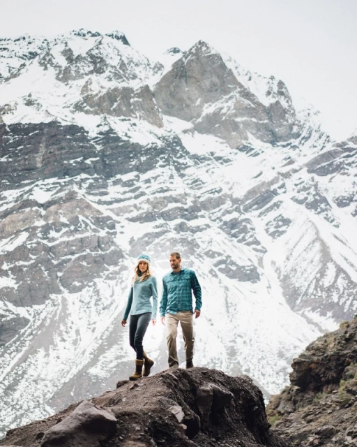 A couple hikes on a rocky path near a large mountain covered in snow. The man wears a plaid shirt, and the woman is in a turquoise sweater.